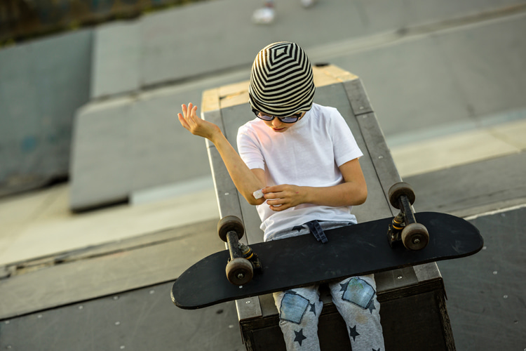 Child wearing a striped hat seated on a skateboard, looking at their arm