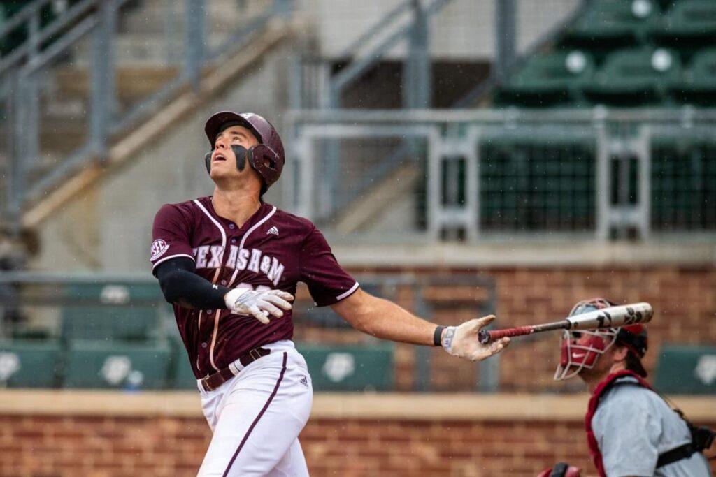 Baseball player in maroon uniform looking up with bat after a swing, catcher behind him