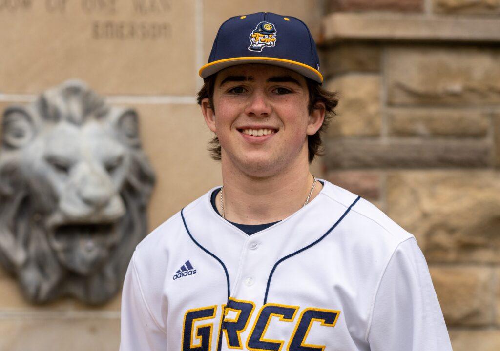 Young man in white baseball uniform and cap smiling in front of a stone wall
