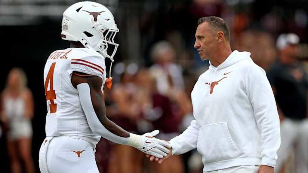 Football player in white uniform shakes hands with coach in white sweatshirt